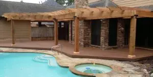 Wood pergola over a backyard patio beside a swimming pool and hot tub.