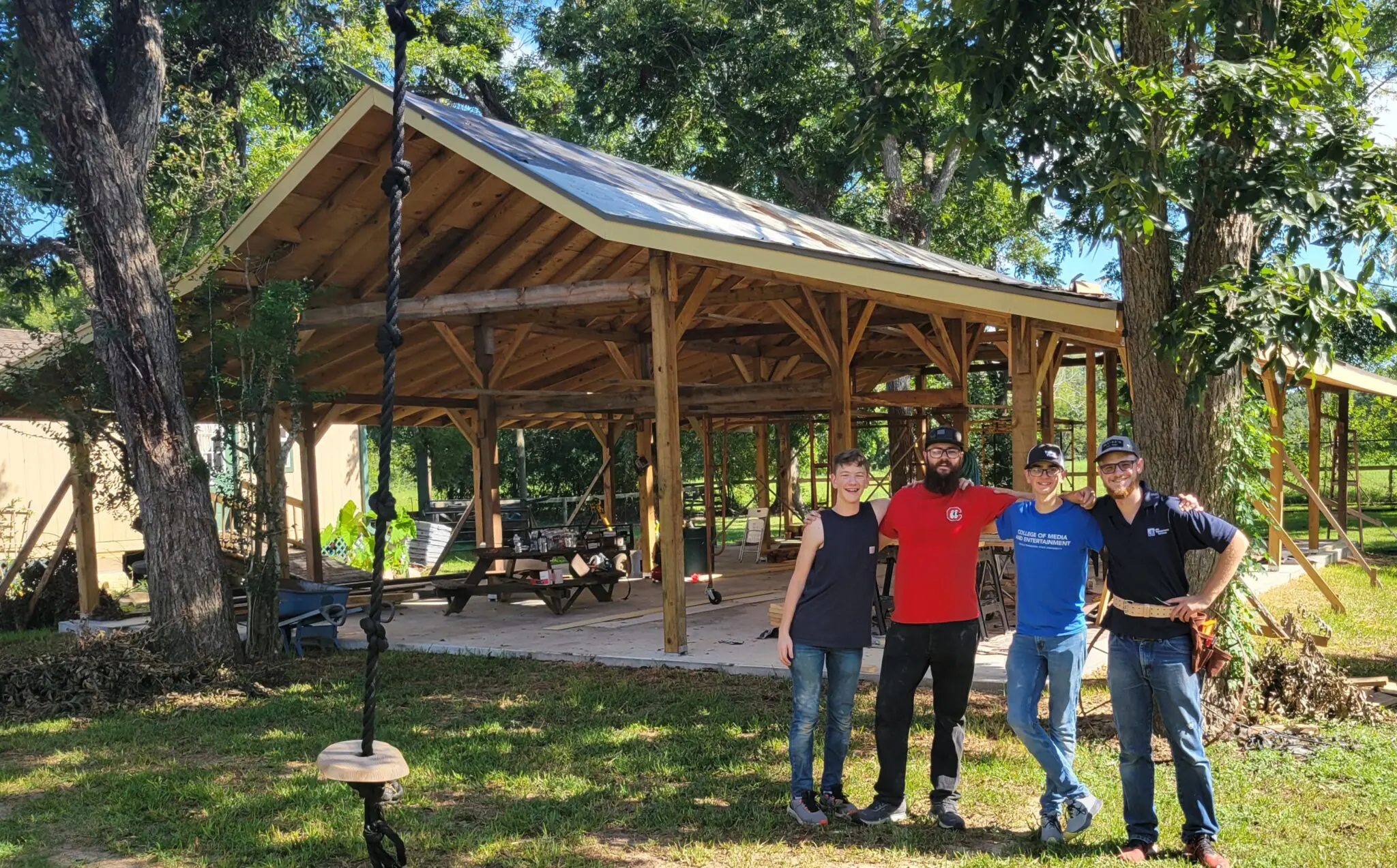 Three men posing in front of a wooden pavilion in a sunny park.