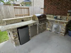 Outdoor kitchen with grill, sink, and refrigerator under a covered patio.