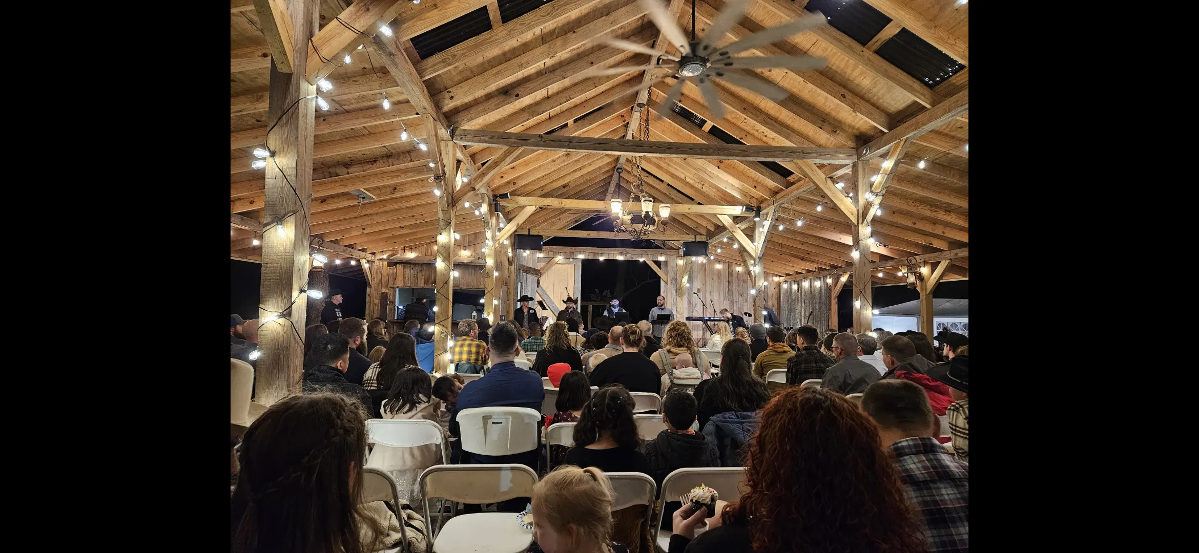 People gathered under a wooden pavilion with string lights for an event.