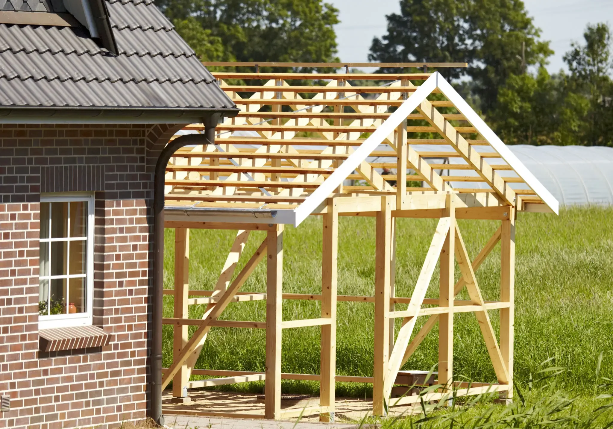 Wooden framework of a greenhouse under construction next to a brick house.